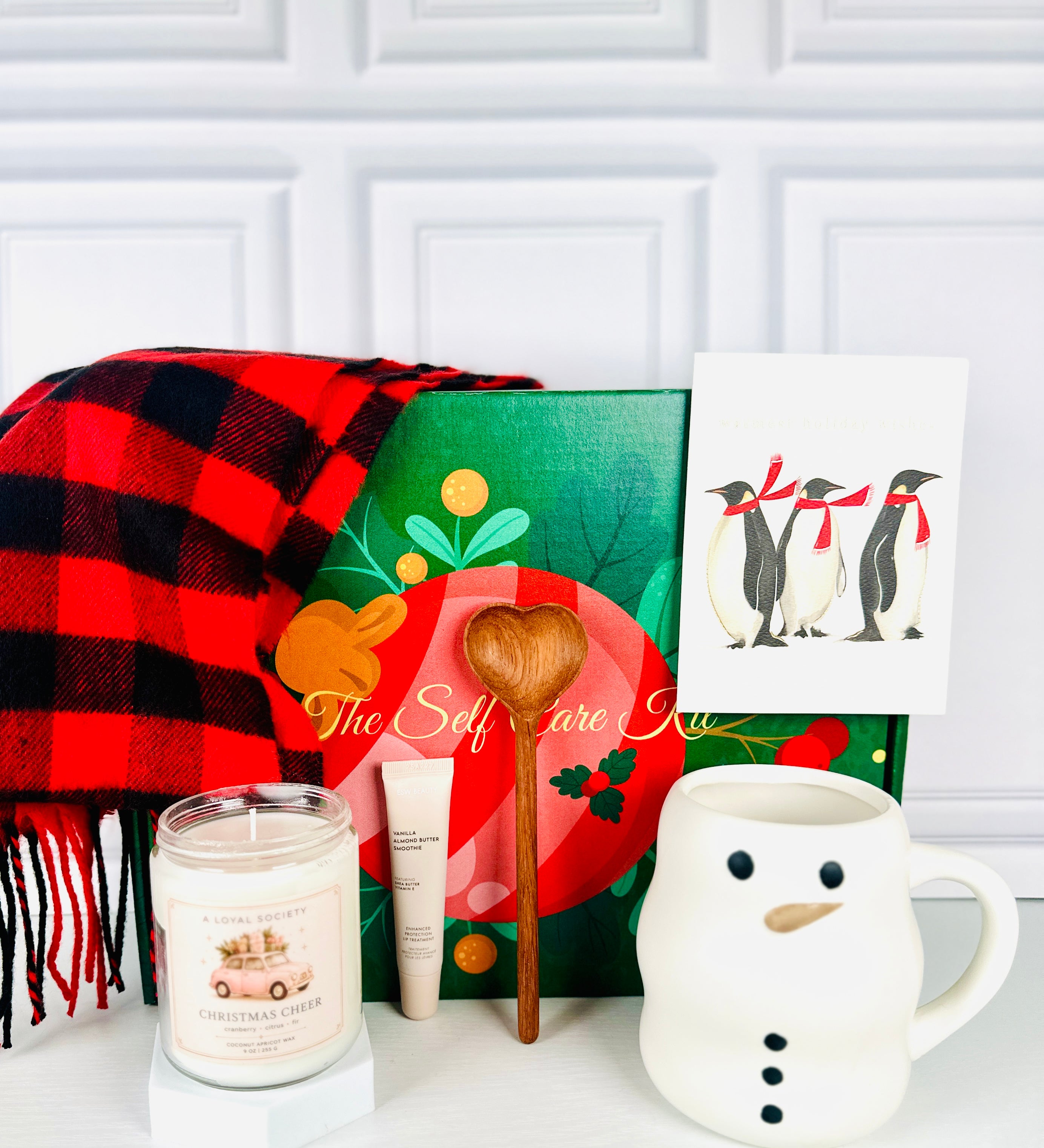 A  Loyal Society Fireside Cozy Christmas holiday gift set designed with a hygge and cozy theme . The giftset features a red and black buffalo plaid scarf, a Christmas Cheer candle, a wooden heart-shaped spoon, a cream- colored snowman mug, lip balm, and a holiday card with penguins wearing red scarves on it. 