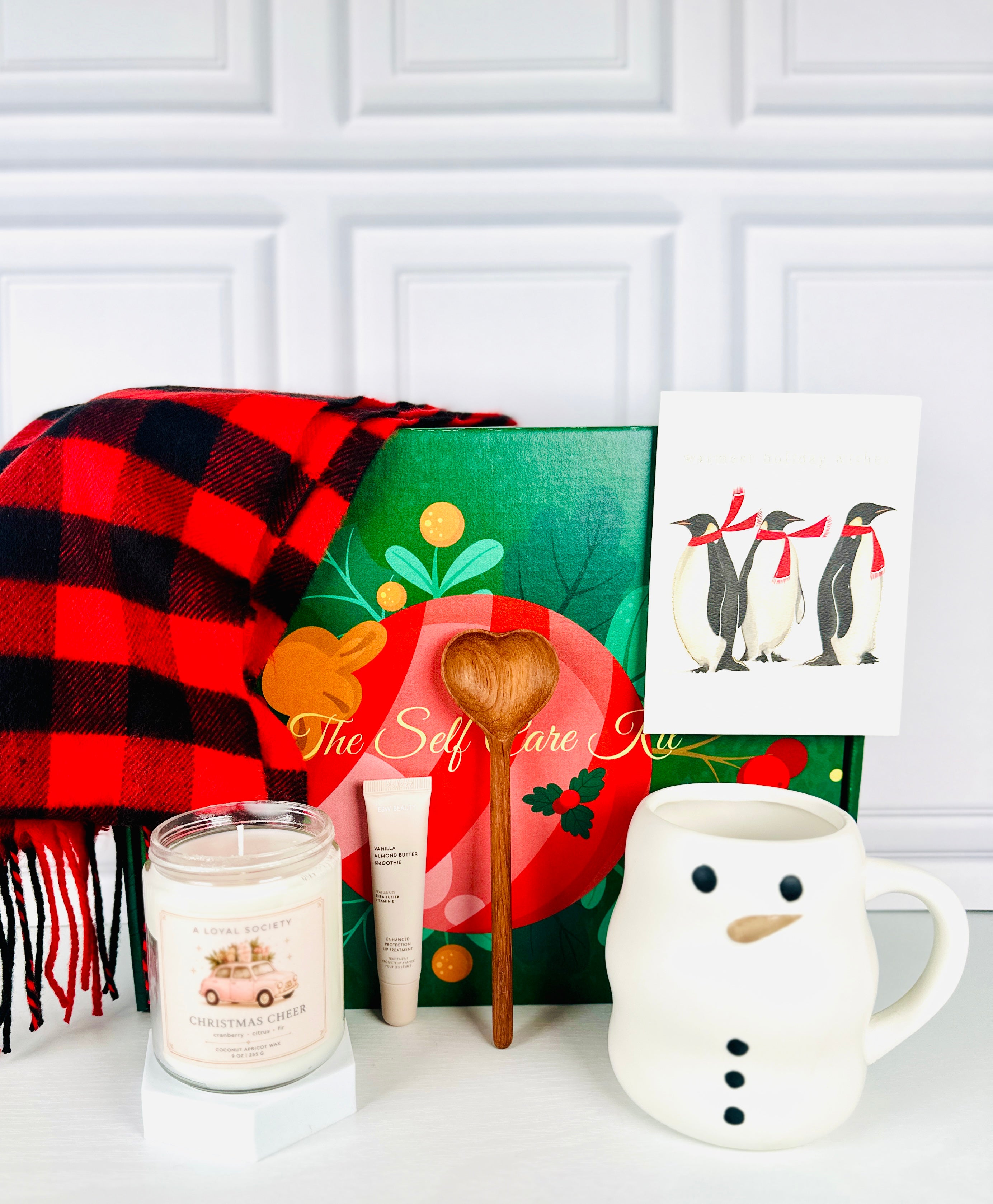 A  Loyal Society Fireside Cozy Christmas holiday gift set designed with a hygge and cozy theme . The giftset features a red and black buffalo plaid scarf, a Christmas Cheer candle, a wooden heart-shaped spoon, a cream- colored snowman mug, lip balm, and a holiday card with penguins wearing red scarves on it. 