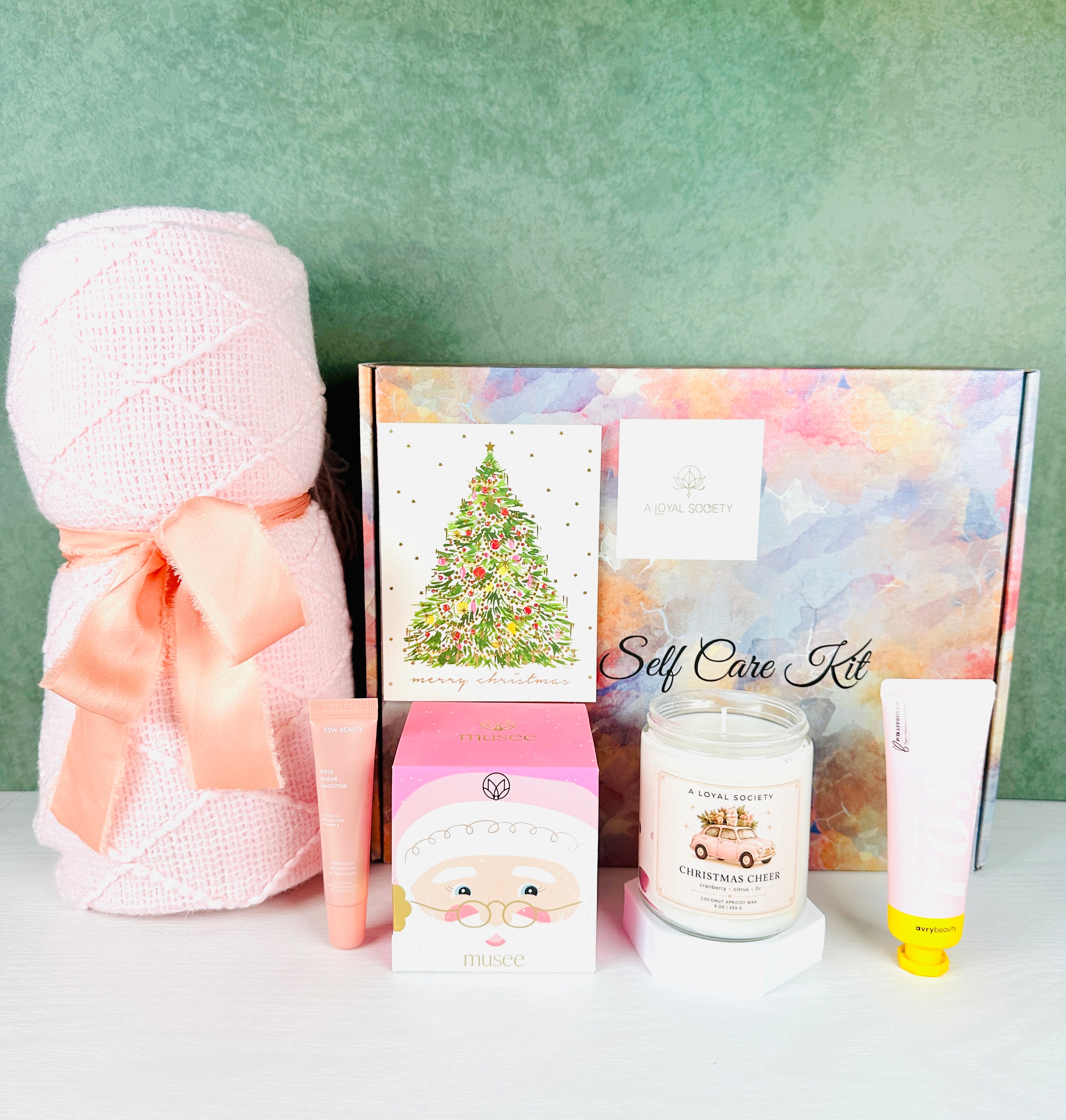 The A Loyal Society Pink Christmas Cheer Spa Gift set is an elegant gift for those who love all things pink. This gift set includes a pale pink knit throw blanket, a pink bath box in a Santa themed box, a Rosewater lotion, a rose lip balm, a Christmas Cheer scented candle and a Christmas tinsel tree card.