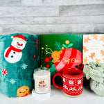 A Christmas-themed self care gift set by A Loyal Society, featuring a plush green blanket with a snowman design, a green and red ornament box with 'The Self Care' branding, a white mug with gingerbread cookies, a candle, and a wrapped gift bag, set against a gray wood plank background.