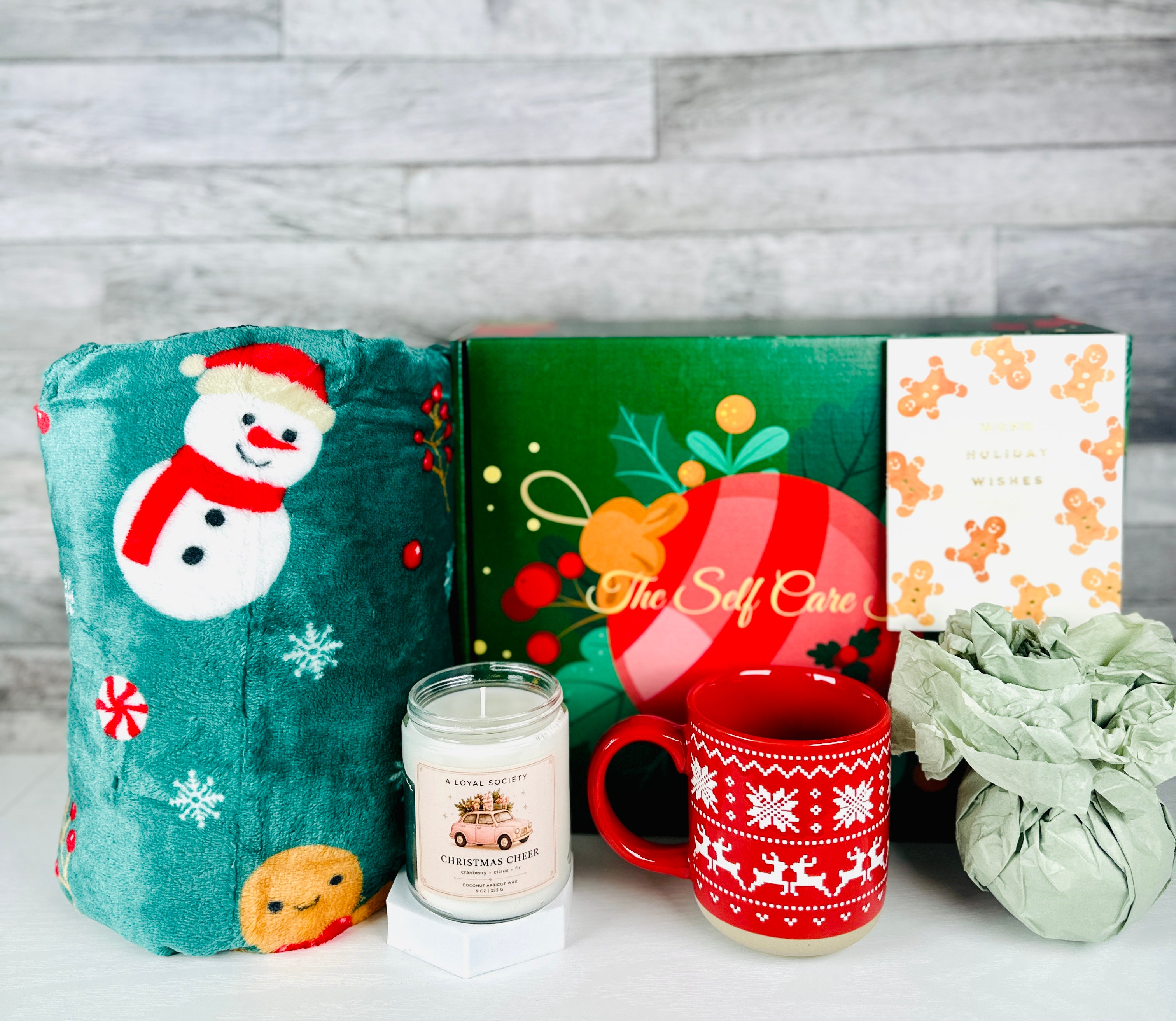 A Christmas-themed self care gift set by A Loyal Society, featuring a plush green blanket with a snowman design, a green and red ornament box with 'The Self Care' branding, a white mug with gingerbread cookies, a candle, and a wrapped gift bag, set against a gray wood plank background.