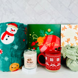 A Christmas-themed self care gift set by A Loyal Society featuring a plush teal blanket with a snowman design, a scented candle, a red mug with a white snowflake pattern, a green gift box, and a wrapped green tissue or paper bundle, arranged on a white surface against a light marble background.