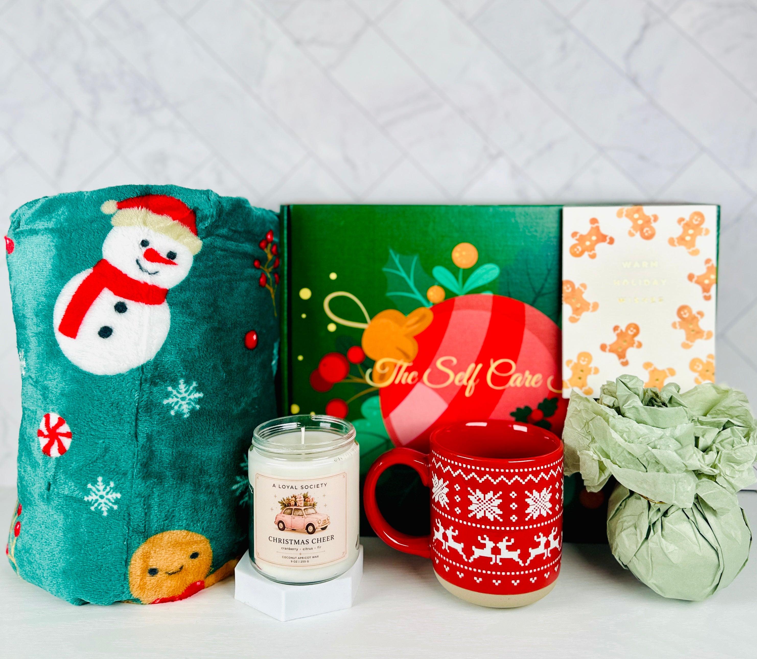 A Christmas-themed self care gift set by A Loyal Society featuring a plush teal blanket with a snowman design, a scented candle, a red mug with a white snowflake pattern, a green gift box, and a wrapped green tissue or paper bundle, arranged on a white surface against a light marble background.