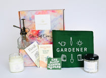 A gift box containing a green apron labeled 'Gardener', a clear plant mister, a relaxation candle, a bath soak, and two packs of non-GMO seeds with 'Spring into Summer' Wildflowers and Basil seeds.